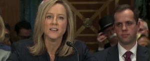 photo of Kathy Kraninger at Senate Hearing with Monopoly Man in background