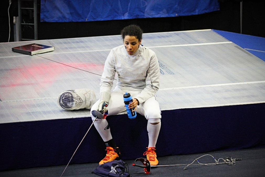 A woman in a fencing uniform sits with her helmet off, holding the fencing sword.