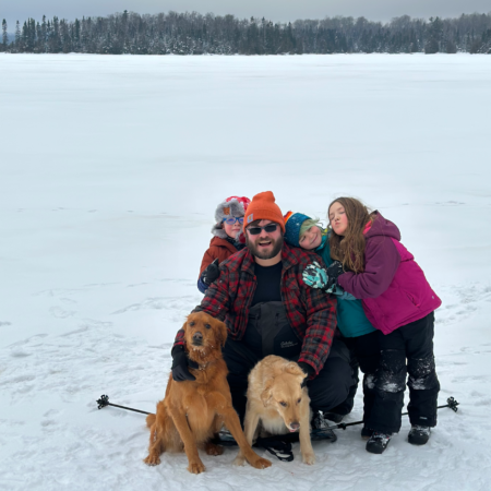 A father with three young children and two golden retrievers in a snowy field.