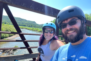 A man and woman standing on a bridge over a river on a sunny day. They are smiling and wearing bike helmets.