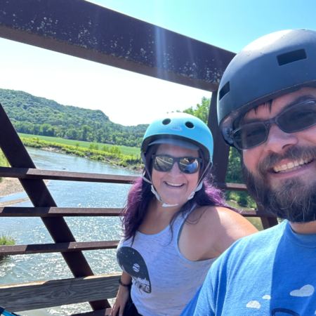 A man and woman standing on a bridge over a river on a sunny day. They are smiling and wearing bike helmets.