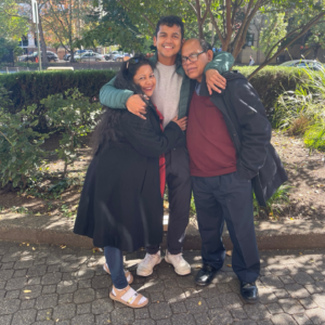 A young adult man hugs his mother and father on a shady sidewalk.