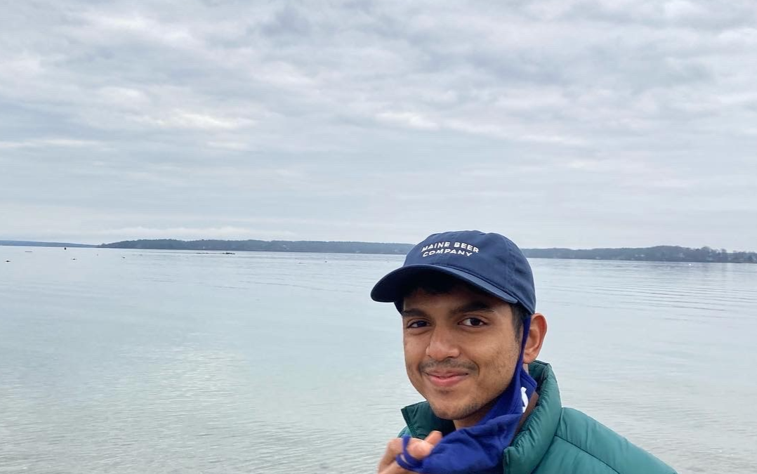 A young man in an insulated jacket and scarf standing in front of a water horizon on a cloudy day.