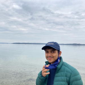 A young man in an insulated jacket and scarf standing in front of a water horizon on a cloudy day.