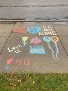 Colorful chalk drawings on a sidewalk, including a birthday cake with candles, balloons, and messages "UR NOT ALONE" and "Love U"