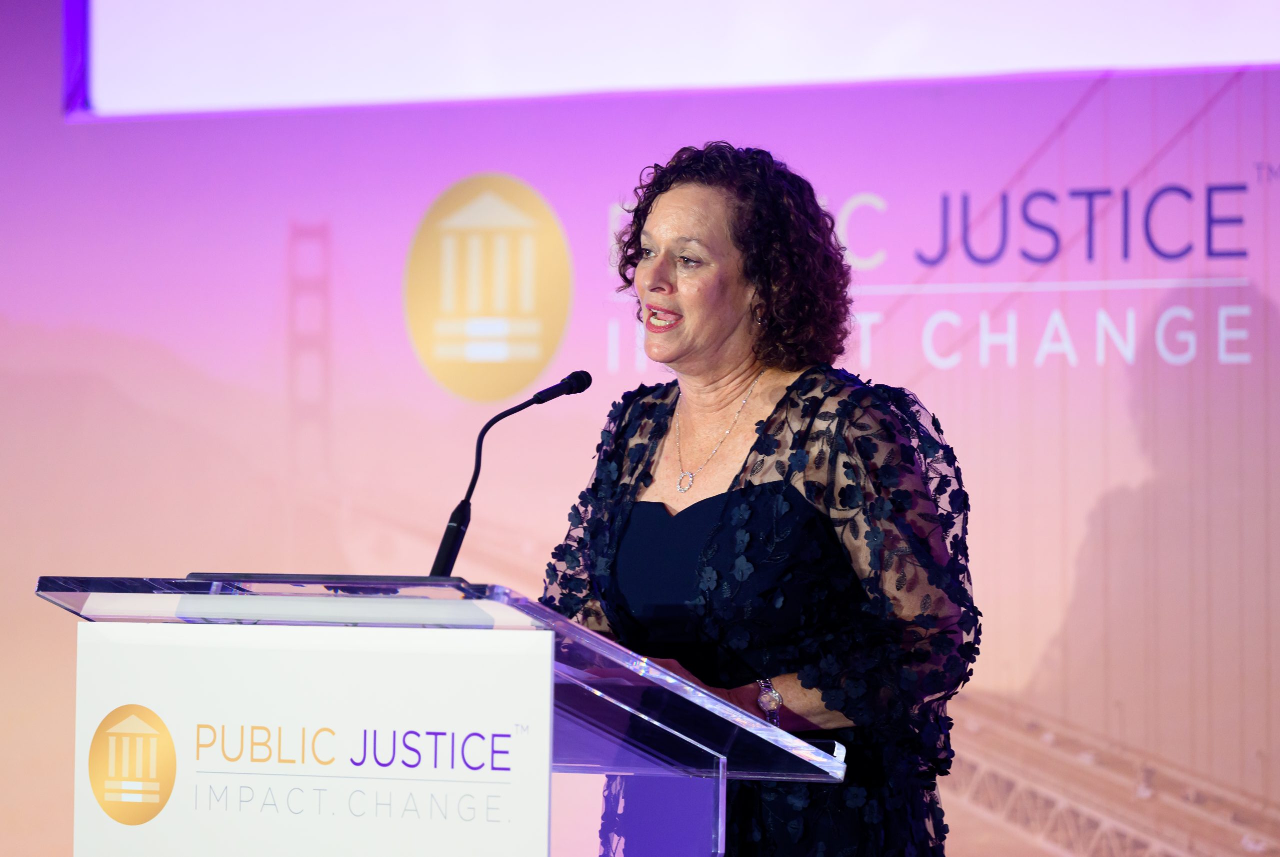 A woman — Public Justice Board President Ellen President — wearing an evening dress stands at a podium giving a speech.